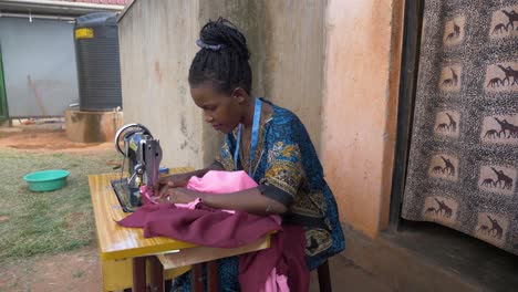 Vista-Lateral-En-Cámara-Lenta-De-Una-Dama-Africana-Cosiendo-Ropa-En-Una-Máquina-De-Sastrería-Manual-Fuera-De-Su-Pequeña-Casa