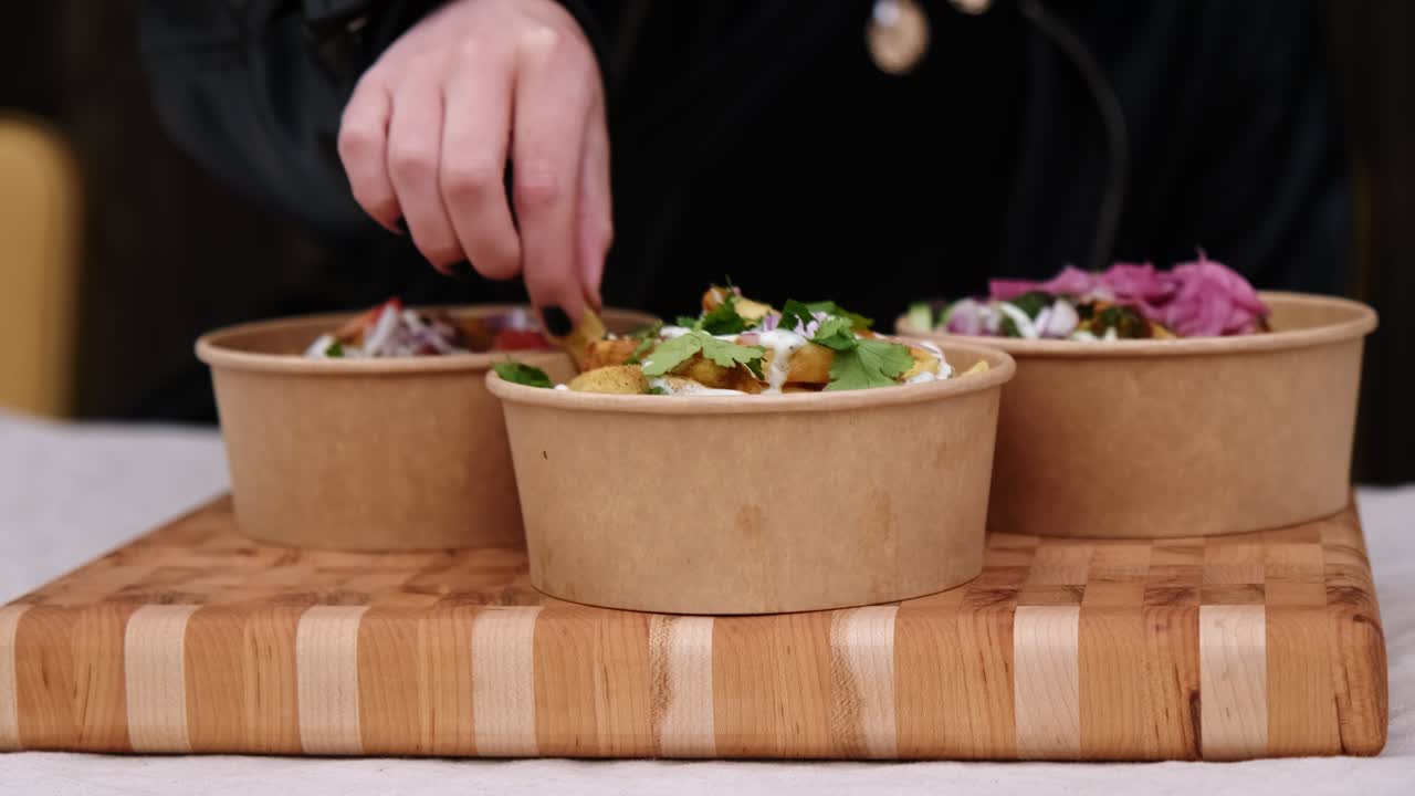 Person eating Indian take away food and fries from biodegradable poke bowl displayed on wooden tray.