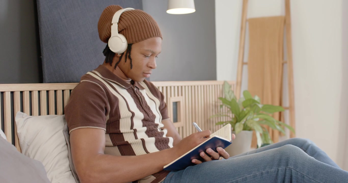 African American man listening with headset sitting bed writing notebook pausing humming at window