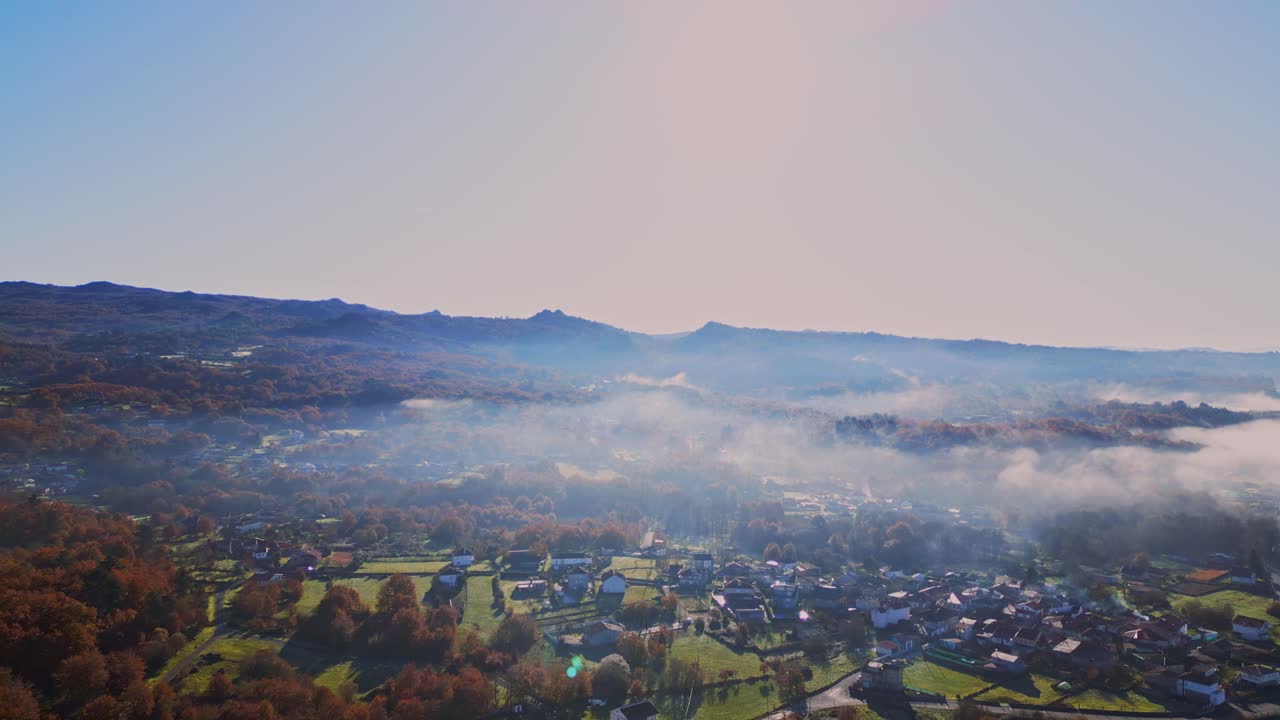 Aerial View Of Mist Covering A Village Surrounded By Forest On Sunny Day