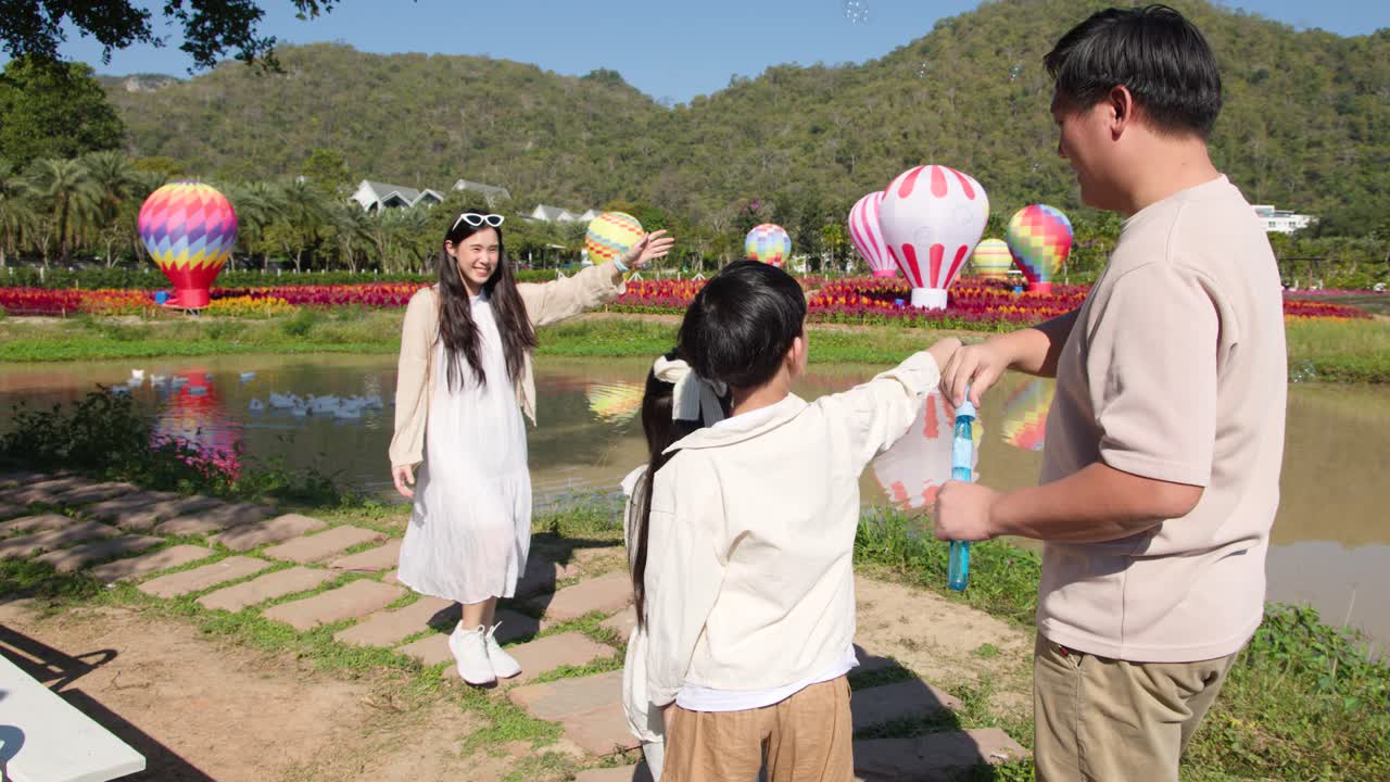 ครอบครัวชาวไทยที่มีความสุขเป่าฟองสบู่อยู่กลางแจ้งใกล้สระน้ำที่มีบอลลูนลมร้อนสีสันสดใส