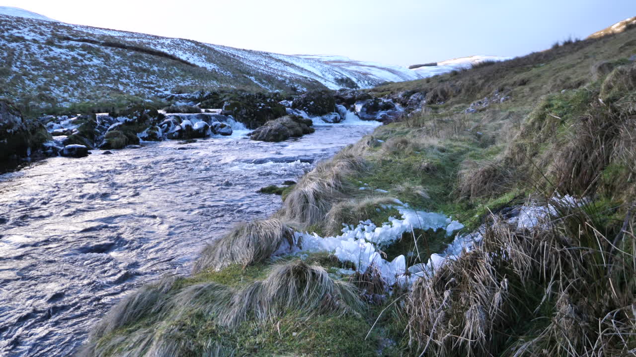 Icy river flowing through snowy hills in Northumberland, UK. Rugged countryside with frozen banks, clear water, and dramatic winter scenery