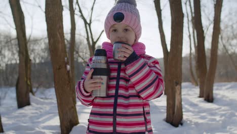 Portrait-of-cute-smiling-child-girl-kid-traveler-walking,-drinking-hot-drink-tea-in-winter-park