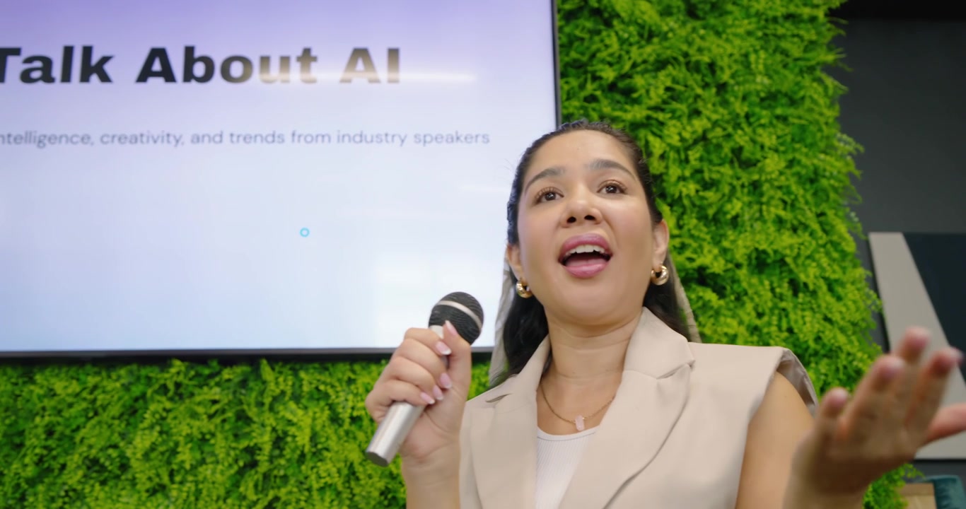 Adult speaker holding mic, explaining, gesturing, pointing to AI slide on stage with moss wall