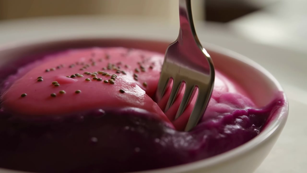 Close-up of a glossy purple dessert in a white bowl as a fork presses into the surface, with small seeds on top and soft natural light against a blurred