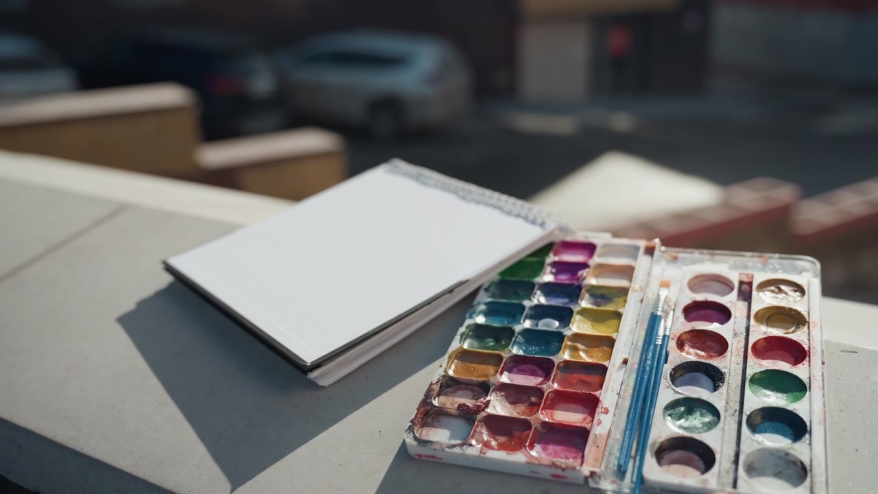 Sonnenbeschienenes Dach-Skizzenbuch mit Aquarellpalette, ordentliche Farbnäpfchen auf dem Sims, weiches Stadt-Bokeh im Hintergrund, subtiler Schatten über dem Papier, einladende kreative und friedliche Atmosphäre zum Malen.