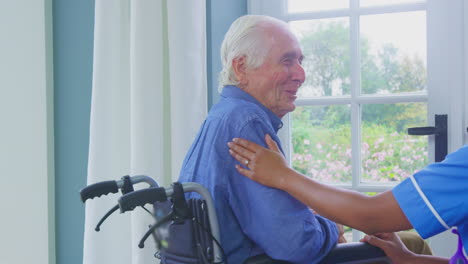 female care worker in uniform talking with senior man sitting in wheelchair in care home lounge