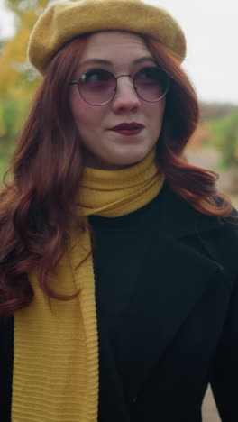 una dama blanca con una boina amarilla y una bufanda a juego ajusta su cabello ondulado rojo mientras hace un paseo de otoño, mirando a su alrededor con un edificio borroso en el fondo lejano