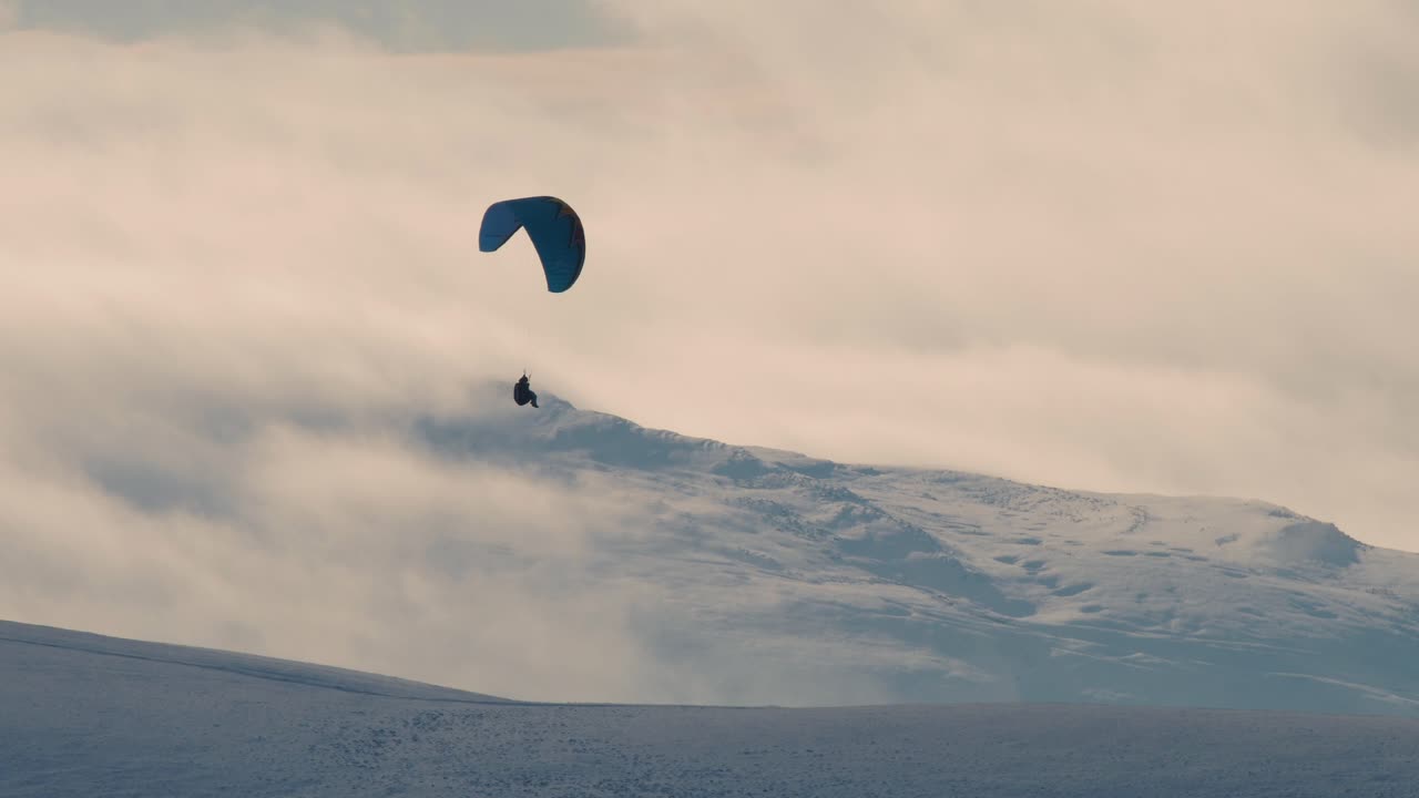 Paraglider shot against the light with snow covered hills and clouds in the background, Cumbria UK