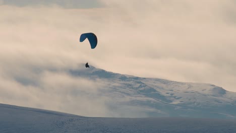Paraglider-shot-against-the-light-with-snow-covered-hills-and-clouds-in-the-background,-Cumbria-UK