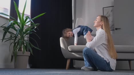 a beautiful young mother is doing yoga and her little daughter is running around the room a beautiful young mother is doing yoga and her little daughter is running around the room fitness classes yoga exercises at home healthy lifestyle