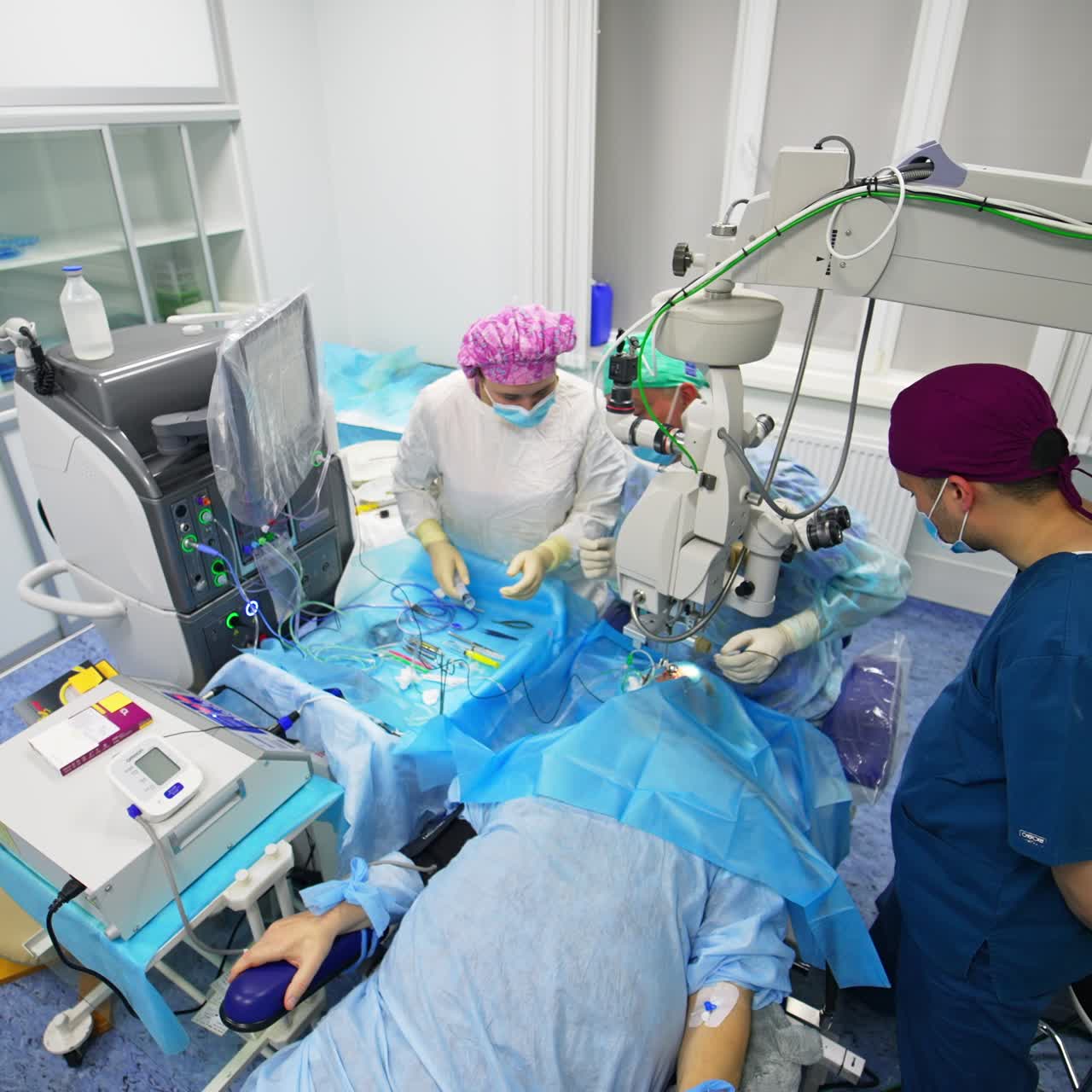 Ophthalmology operation performed at the well-equipped surgery room. Patient lying on the table under the modern equipment. Surgeons look into microscopes