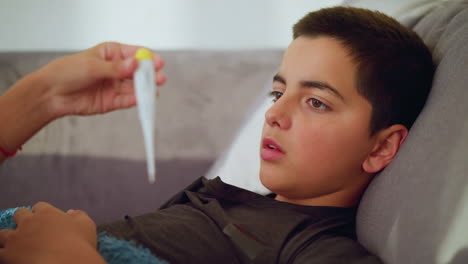 close up of sick boy lying on couch under blanket looking at thermometer held by another person showing concern and awareness in calm indoor space