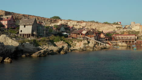 Aerial-Establishing-Shot-of-Popeye-Village-in-Anchor-Bay,-Gozo-Island,-Malta-wooden-Village-Houses-by-Water,-Aerial-Drone-Shot-forward-Dolly