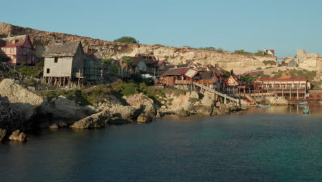 Toma-Aérea-Del-Pueblo-De-Popeye-En-La-Bahía-De-Ancla,-Isla-De-Gozo,-Casas-De-Pueblo-De-Madera-De-Malta-Junto-Al-Agua,-Drone-Aéreo-Disparado-Hacia-Adelante-Dolly