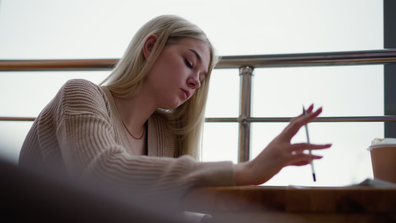 Contemplative lady pauses as she hits pen on table, continues writing, blur of coffee cup and iron rail in background with urban setting visible outside window