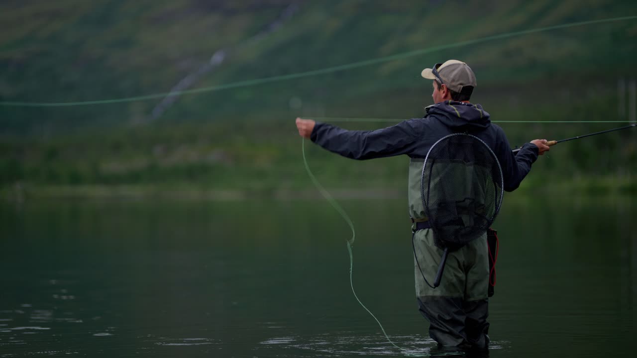 Young man casts fly fishing rod back and forth at calm lake, side view