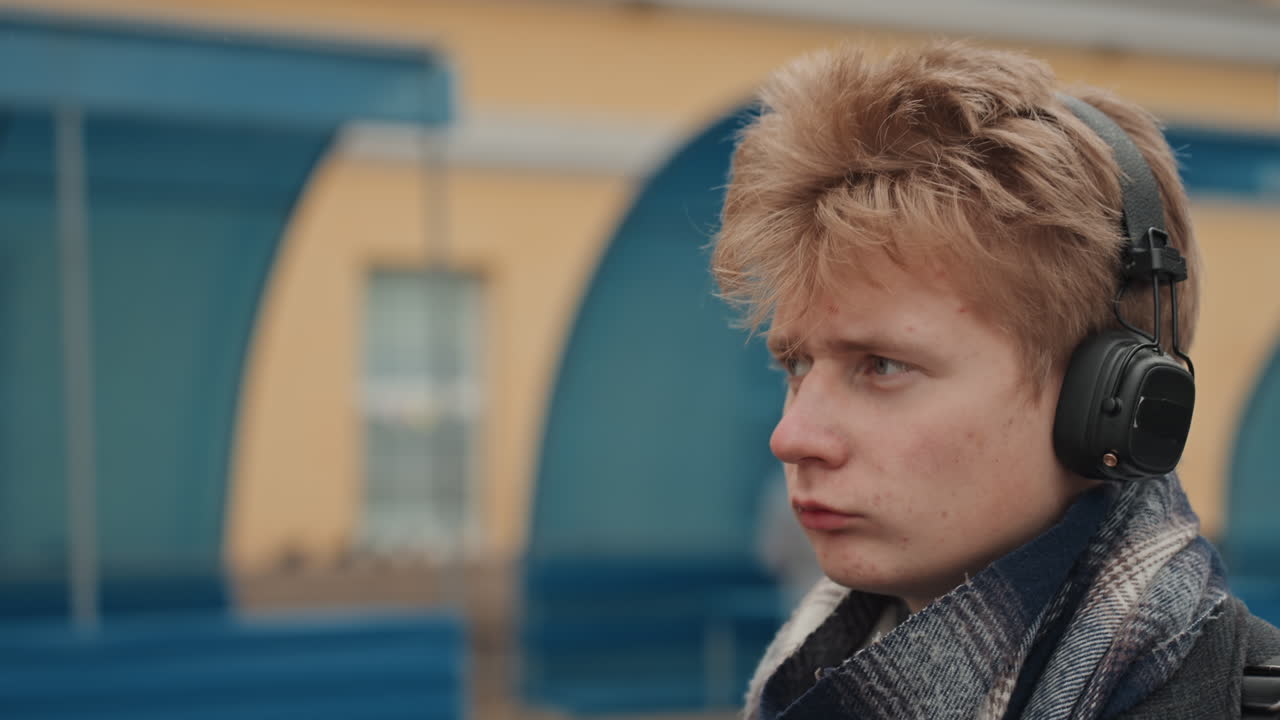 Closeup white creator listening with headphones at bus shelter, winter breath and focused stare, blue shelter backdrop, pensive mood, freeze on hair and scarf, quiet urban waiting scene that feels