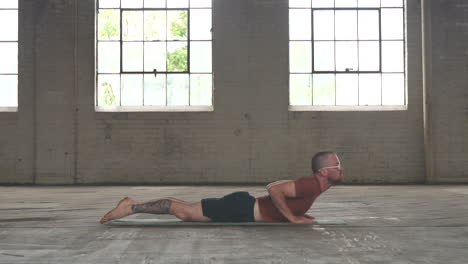 man in an industrial warehouse practicing yoga doing a cobra pose and moving into a plank pose.