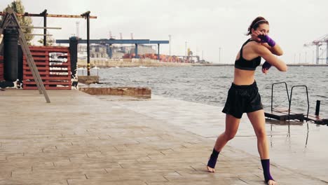 Mujer-Joven-Atlética-Haciendo-Boxeo-De-Sombras-Junto-Al-Mar.-Hermosa-Boxeadora-Entrenando-En-La-Playa-Por-La-Mañana,-Lanzando-Golpes