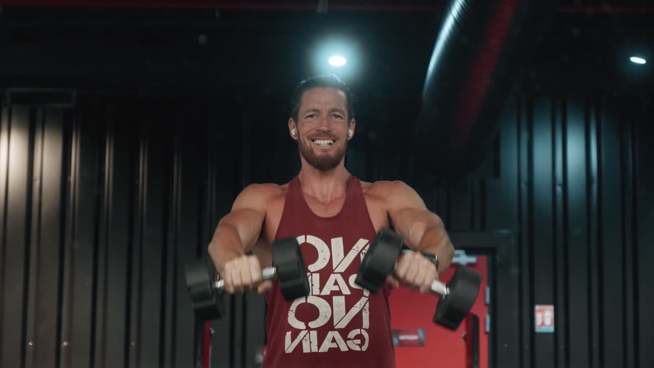 Focused male athlete lifting dumbbells in the gym, wearing a motivational “No Pain No Gain” tank top. Strength, discipline, and determination in action