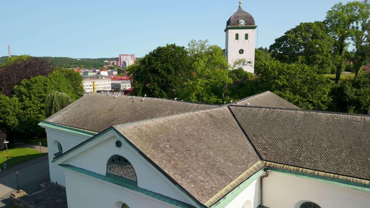 Letecký paralaxní záběr dronem obíhající bílou neoklasicistní střechu kostela Uddevalla se zvonicí, hodinami a siluetou města viditelnou v pozadí