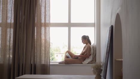 lady student writes graduate work via laptop on windowsill