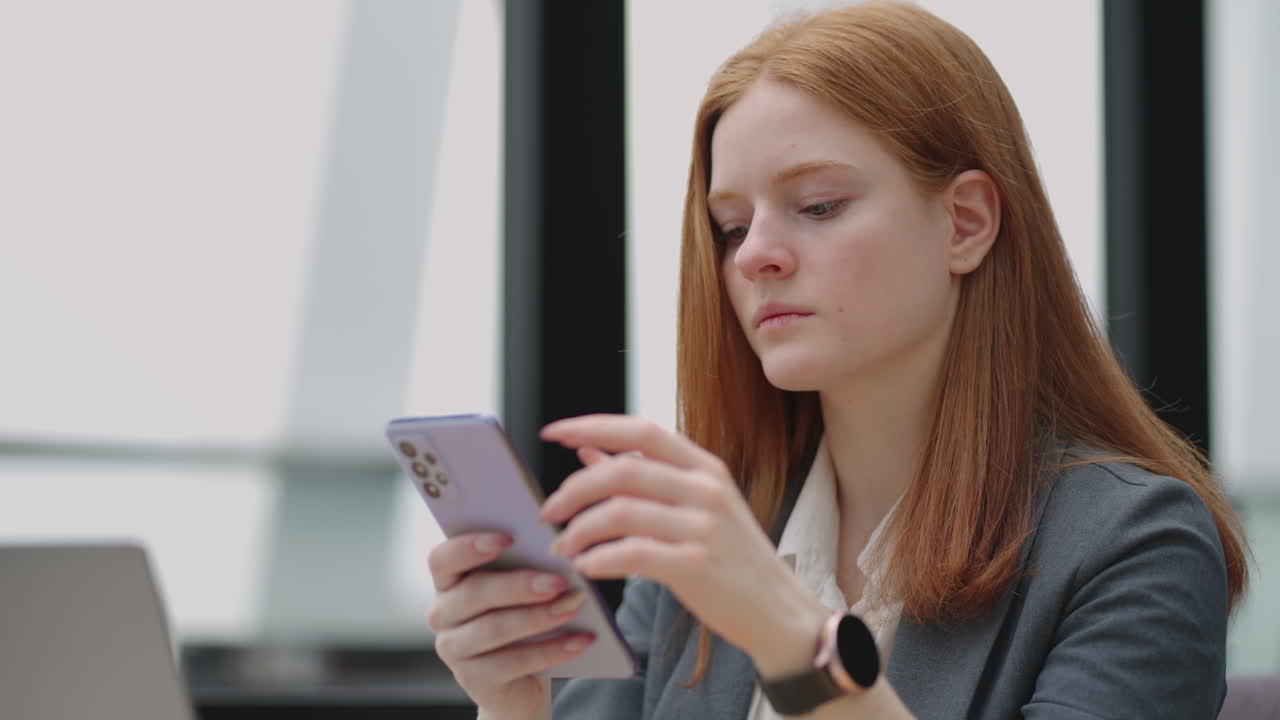 Beautiful young business woman using phone in office. Woman using a smartphone and leaning on a window texting sending emails planning meetings networking online browsing messages on mobile phone