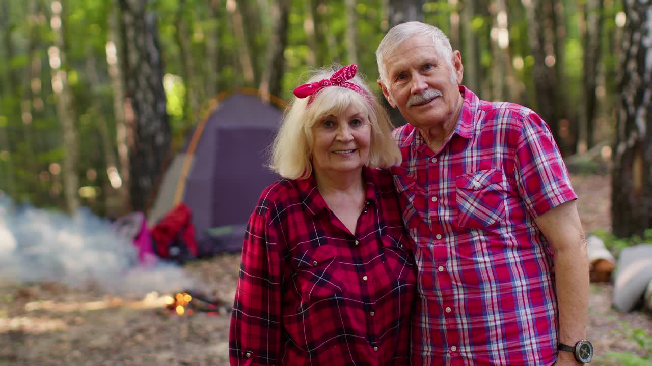 Portrait of old happy couple resting at camping in forest over campfire and tent background