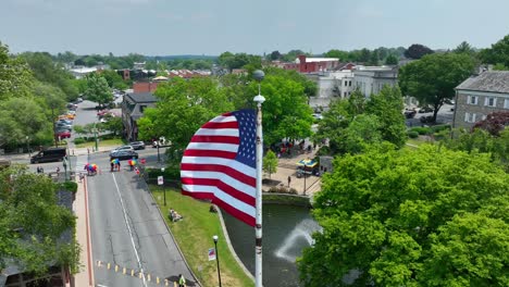 Drone-view-of-a-city-preparing-for-a-pride-festival,-people-gathering-near-a-park-and-the-American-flag-waving-in-the-breeze-on-a-sunny-summer-day