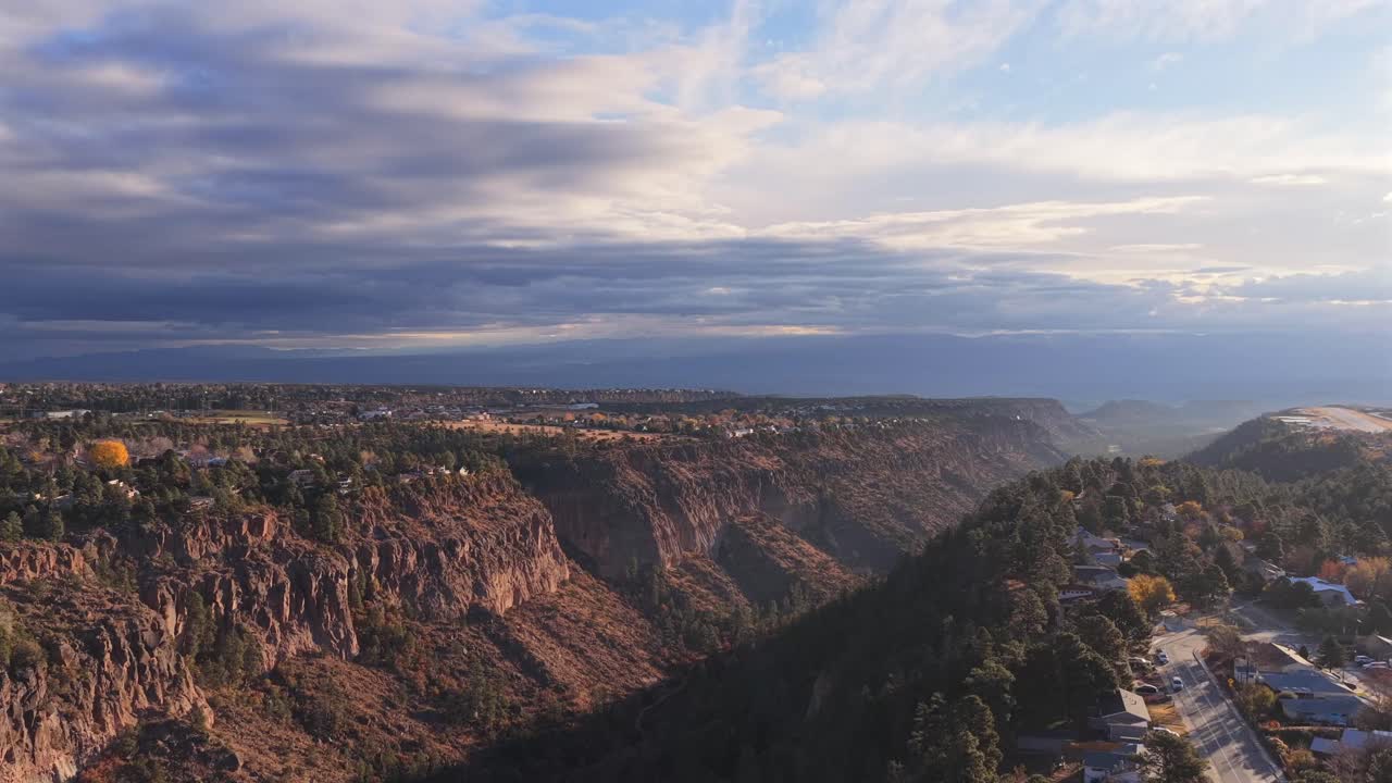 Wide aerial shot featuring residential town development on a forested mesa top. The Los Alamos airstrip is visible in the background, set against the steep, dramatic canyon gorge at sunrise