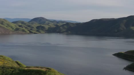 Drone-shot-orbiting-beautiful-mountain-top-covered-in-yellow-wildflowers,-green-grass-and-large-lake-reservoir-during-the-California-super-bloom