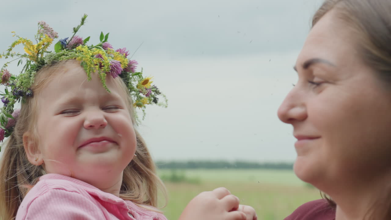 Madre e hija, profesoras blancas, intercambian una mirada cariñosa en un prado; la niña, que lleva una corona de flores, sonríe; perfil reflexivo de la madre; paisaje rural tranquilo; recuerdo nostálgico de verano; vínculo afectuoso.