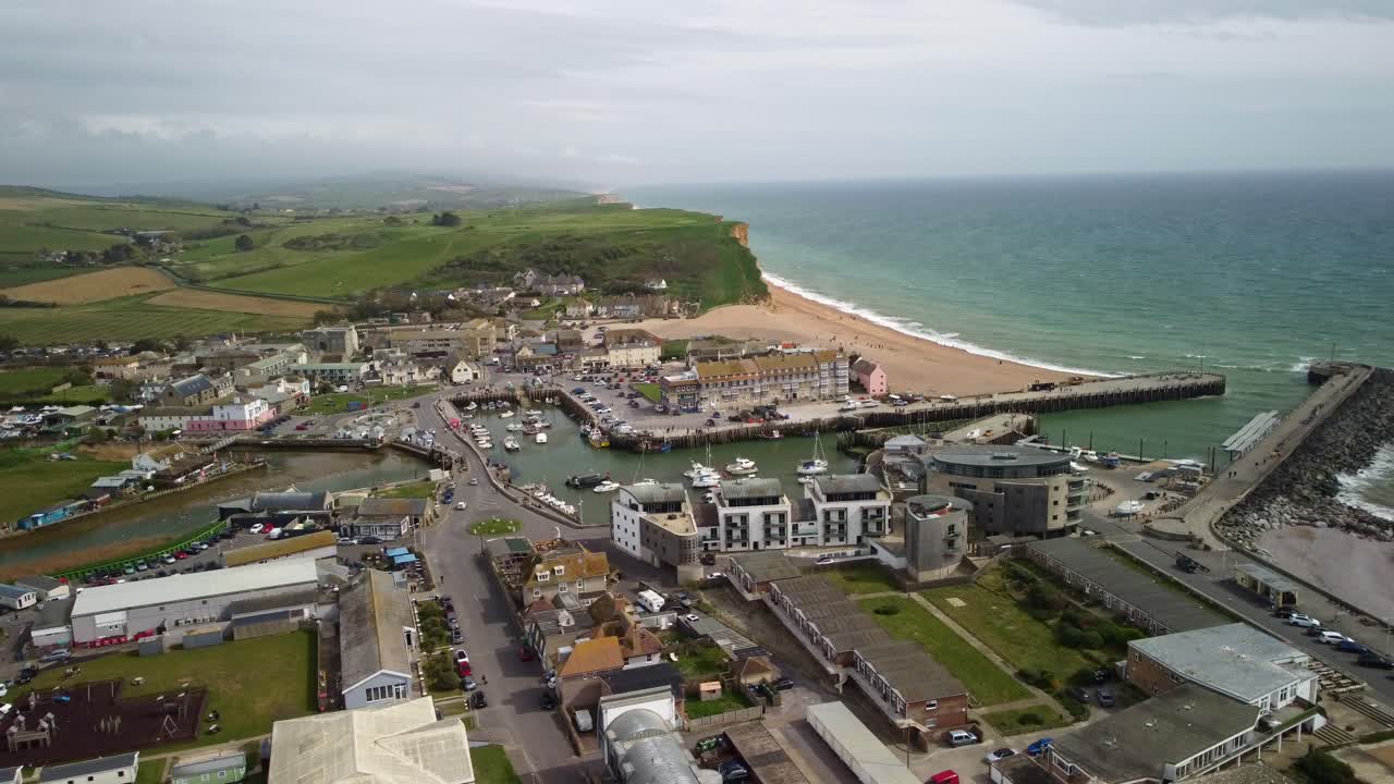 Aerial footage moving sides over West Bay, Dorset. The drone moves from the harbour centre to the right out towards the sea. shops, buildings, boats, cars, the beach, Jurassic Coast cliffs.