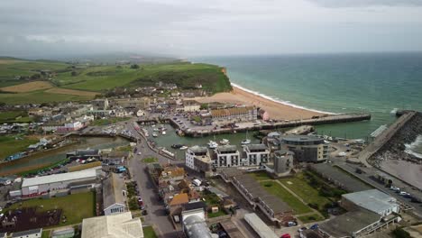 aerial footage moving sides over west bay, dorset. the drone moves from the harbour centre to the right out towards the sea. shops, buildings, boats, cars, the beach, jurassic coast cliffs.