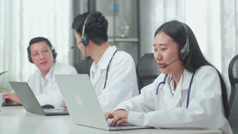 a woman of three asian doctors with stethoscope in headset working as call center agents speaking to customers on the call and typing while her colleagues are talking during working at the office