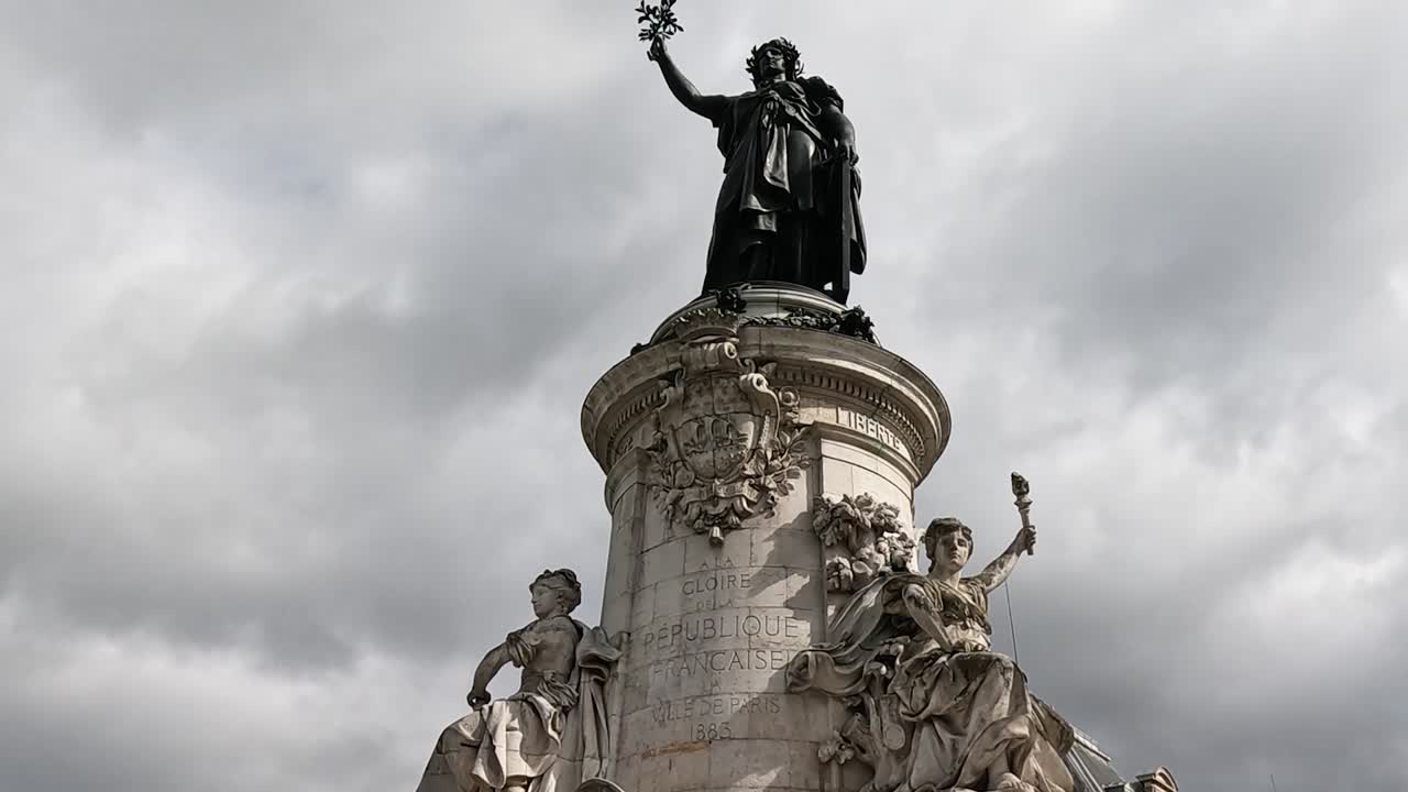 A towering statue on a pedestal with intricate sculptures, set against a dramatic cloudy sky.