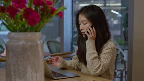 Young-Asian-girl-talks-actively-on-the-phone-in-front-of-her-laptop,-while-working-remotely-from-coffee-shop