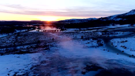 panoramic view with a drone of a geyser in geysir, iceland. smoke is getting out of the geyser with people walking all around the geyser, under the red and orange sunset.