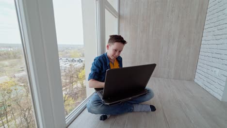 teen boy plays on the gadget on a balcony. boy sitting by the window and playing video games on a modern laptop expresses different emotions.