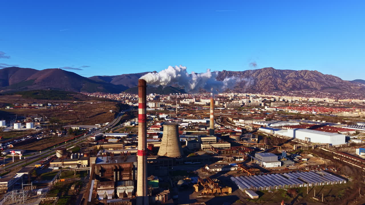 Fumaça sobe de torres industriais enquanto trabalhadores realizam suas tarefas em uma área de fabricação sob um céu azul claro em uma região montanhosa.