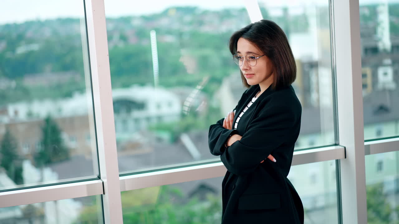 Beautiful brunette woman wearing glasses and black jacket stands at the window. Portrait of a confident lady in office. Blurred backdrop.