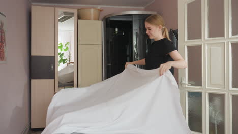 young woman in black outfit spreads white bedsheet over bed in softly lit room with wall mirror and organized shelves in background, preparing space in calm, clean, and minimal indoor setting