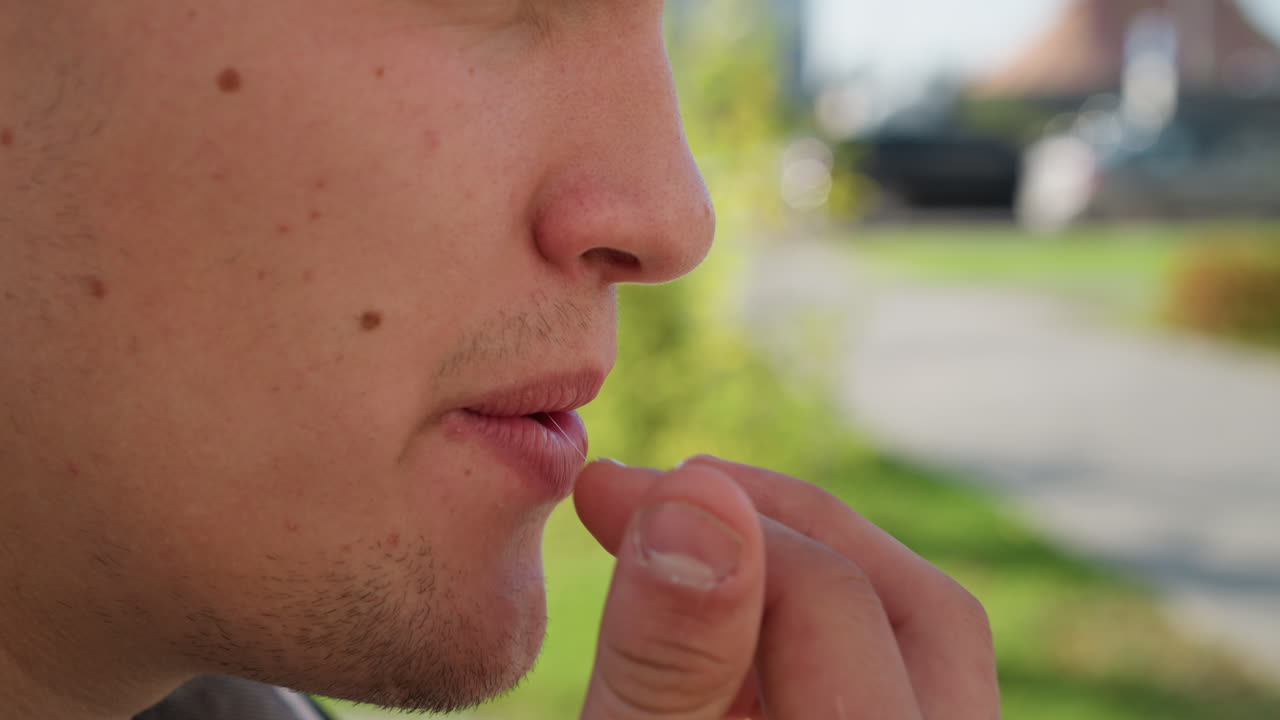 extreme close up of student placing sweet in mouth with natural sunlight highlighting facial features and lips as blurred green background and distant urban elements blend into soft bokeh