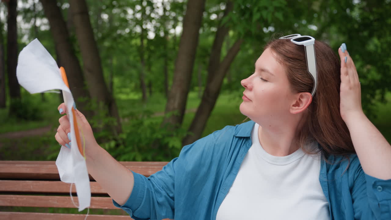 White girl sitting on park bench holding embroidery hoop toward sunlight, observing fabric as if mirror, surrounded by tall green trees, calm natural park atmosphere