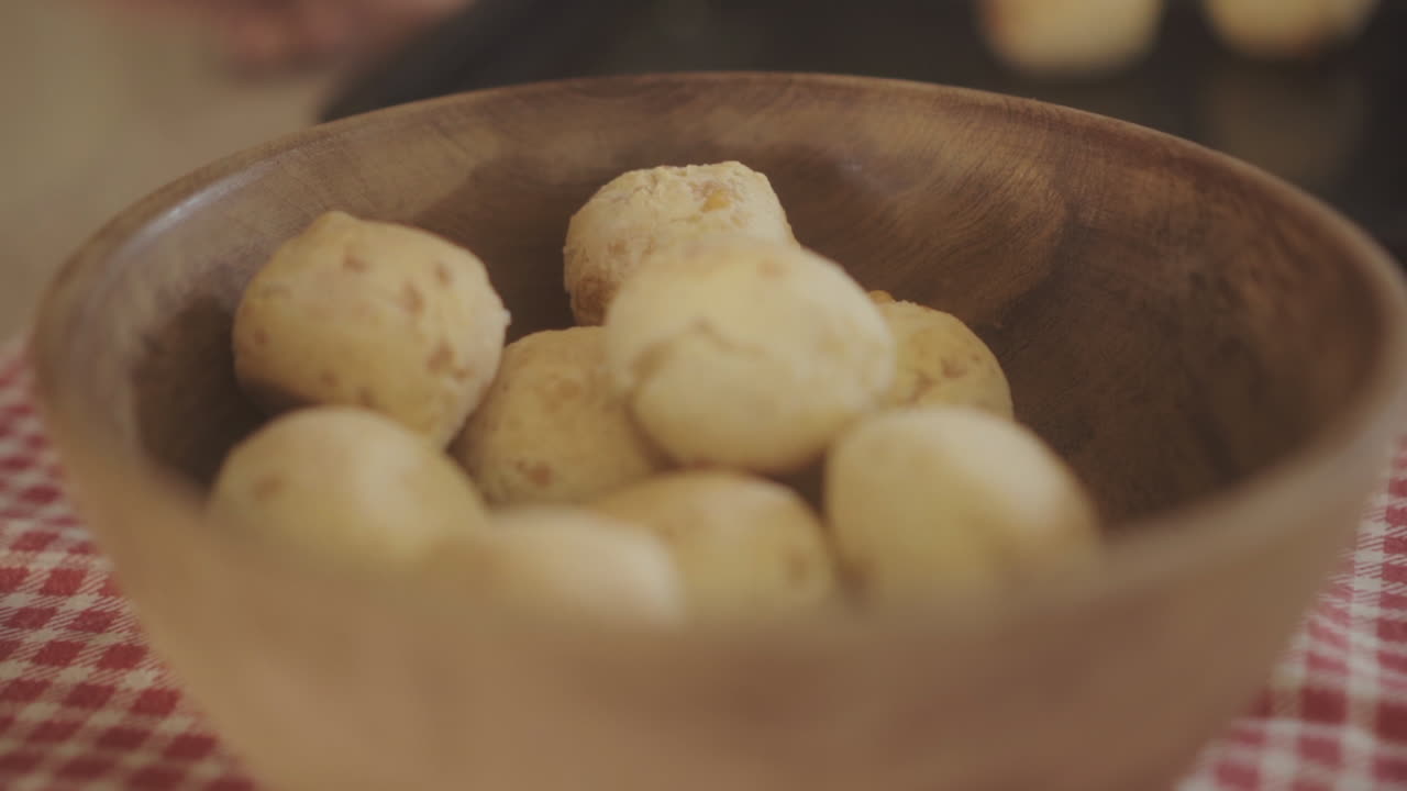 las imágenes de cerca muestran a una persona poniendo panes de chipa recién horneados en un cuenco de madera rústico, colocado en una tela a cuadros roja y blanca, capturando la esencia de la cocina tradicional casera