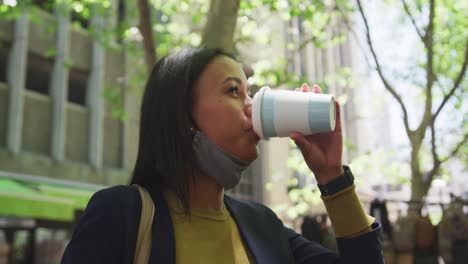 African-american-woman-wearing-face-mask-drinking-coffee-in-city-park