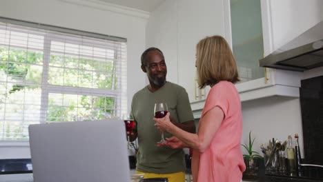 Diverse-senior-couple-in-kitchen-using-laptop-and-drinking-wine