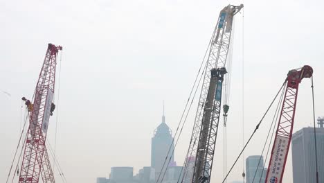 Zahlreiche-Kräne-Stehen-Und-Arbeiten-Auf-Einer-Baustelle-Im-Rahmen-Eines-Gewerblichen-Sanierungsprojekts,-Während-Die-Skyline-Des-Finanzviertels-Von-Hongkong-Im-Hintergrund-Zu-Sehen-Ist