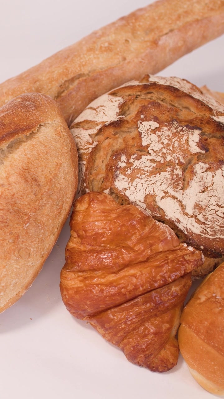 Close-up slow panning shot of sourdough, baguettes, croissants, and sliced grains on white surface.